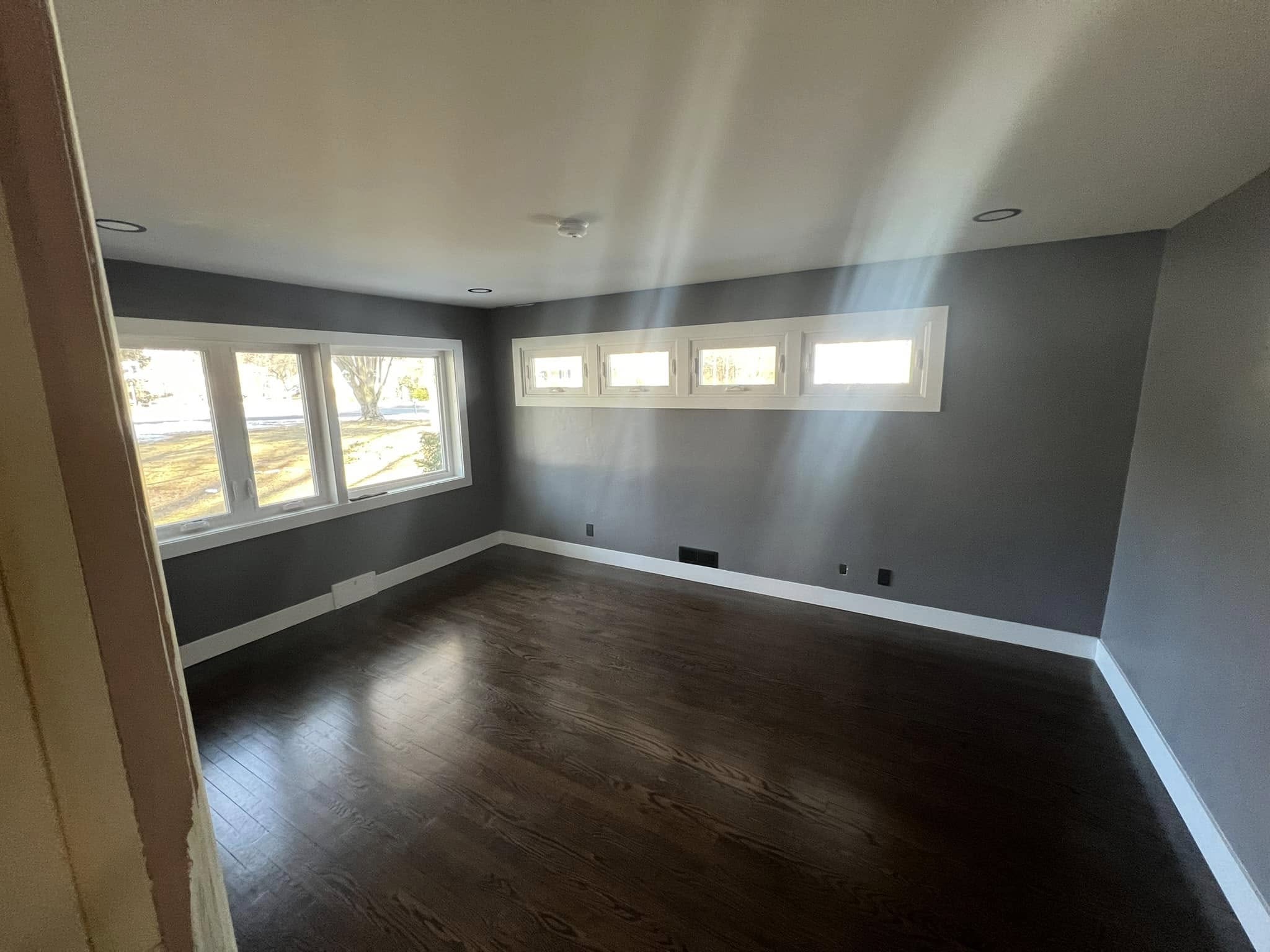 Freshly added room with dark hardwood flooring, smooth gray walls, and bright white trim. Features a row of large windows on one wall and smaller rectangular transom windows on another, allowing natural light to fill the space. Located in Windsor, Connecticut.