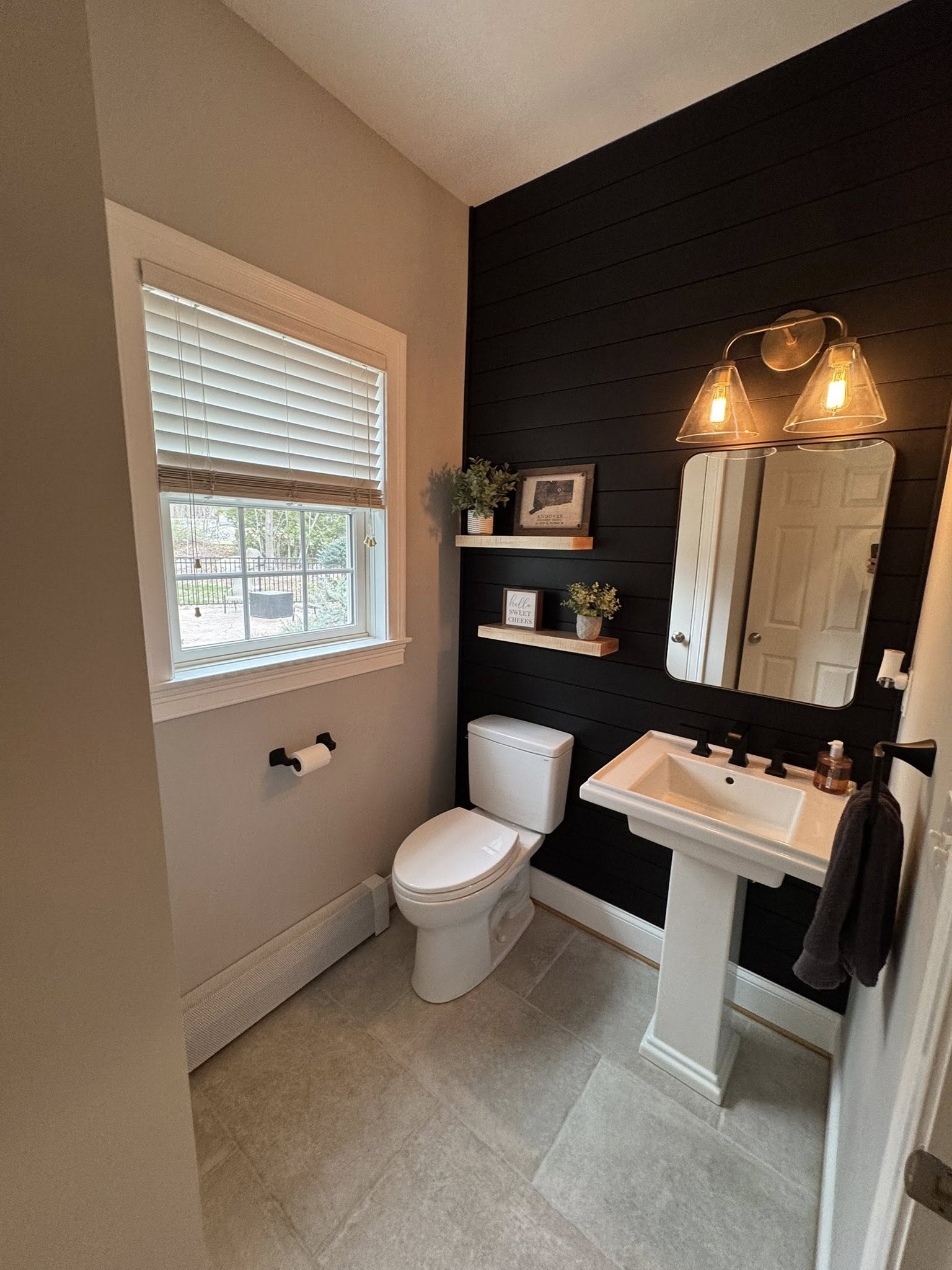 Modern half bathroom remodel in Hebron CT featuring a black shiplap accent wall, pedestal sink, white toilet, wall-mounted shelves with décor, and warm lighting above the mirror.